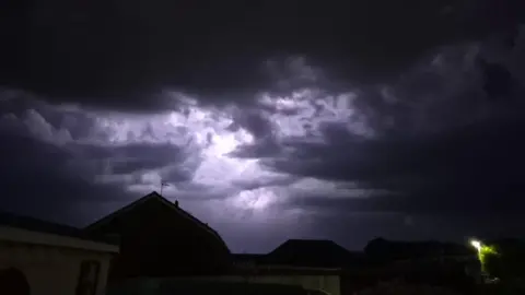 Weather Watchers/McJunior The sky over Whitchurch in north Somerset. There are dark clouds and a flash of white in amongst them from a lightning bolt. The silhouettes of houses can be seen at the bottom of the frame.