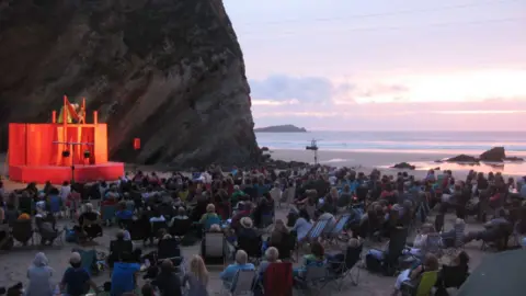 Miracle Theatre A view of a theatre stage situated on a beach with a crowd of people looking towards the stage. The stage is lit up and to the right is a sea-view as the sun sets.