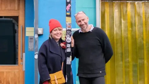 Cobalt Studios Kate Hodgkinson and Mark Collet standing in front of Cobalt which is a blue building with a yellow entrance. They are standing by a black pole which has various stickers on it. Hodgkinson is on the left and is wearing an orange hat with a blue coat and yellow bag across her body. Collet is on the right and is wearing a dark jumper over a shirt and has glasses on top of his head. They are both smiling into the camera. 