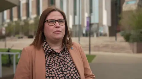 A woman in a brown jumper. She is wearing glasses and has straightened brown hair. 