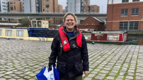 Mark Ansell / BBC Naomi Roberts from the Canal & River Trust smiling at the camera on the cobbled towpath next to Sheffield and Tinsley Canal. She has blonde tied-back hair and is wearing glasses and a black waterproof coat and carrying a blue and white umbrella. In the background are two narrowboats moored alongside the path.