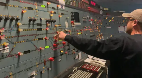 GTR The inside of a signalling centre, with grey/blue panels. There are lots of knobs and buttons. some lit up, and a rail map on the walls. A man wearing a beige baseball cap and black sweater stands with his back to the camera and twiddles a red knob. 