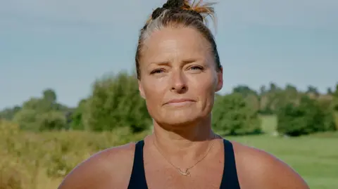 Ellie Ramsden A head and shoulders photo of Laura Reineke, a woman with blonde hair tied in a bun. She's wearing a black swimming costume and a necklace with a mermaid on. She's looking straight at the camera with a half-smile, with trees and a blue sky behind her.