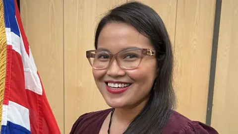 A woman from The Philippines wearing glasses and a burgundy dress stands next to a picture of the King which is draped by a Union Jack