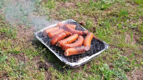 Sausages sizzling on a disposable barbecue. The barbecue is on a patch of grass.