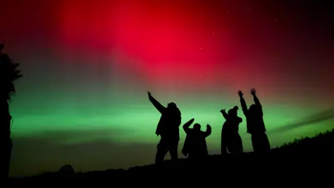 Silhouetted figures raise their arms toward the sky beneath a striking display of red and green aurora lighting the night.