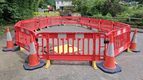 A large circular arrangement of cones and bollards blocks a quiet leafy road. 