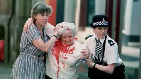 PA Media A female police officer and an unidentified woman help a woman wearing a blood-stained shirt in the aftermath of the IRA Manchester bomb in June 1996.