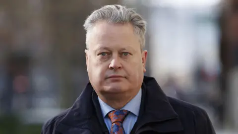PA Media Vincent Kearney is looking directly at the camera on an outside street. The background to the image is out of focus. Vincent has short grey hair and is wearing a navy trenchcoat, blue shirt and colourful red tie, which is covered in blue flowers. 