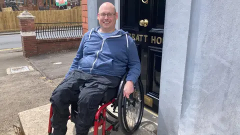 A man with a shaved head and glasses is sitting in a red wheelchair outside a door, smiling. He is wearing a blue hoodie and black trousers.