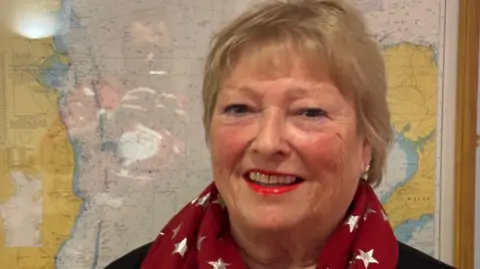 Oli Dimelow Gilli Cowley, pictured with short blonde hair, wearing red lipstick and a red scarf with a map in the background