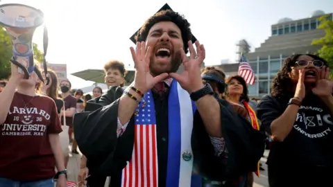 A Harvard University student chants with fellow demonstrators during a protest in support of international students at Harvard University.  He is wearing a graduation cap and gown with a sash which has an American flag on it. 