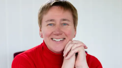 Diane Danzebrink is smiling at the camera while wearing a red turtleneck top and silver stud earrings. She has light brown short hair and blue eyes. She has her elbows on the table and hands clasped upward in front of her, so her chin rests on them.