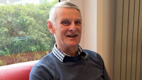 A man in his 50s sat on a red leather sofa. He is smiling, wearing a nave blue jumper and a blue and white check shirt. 