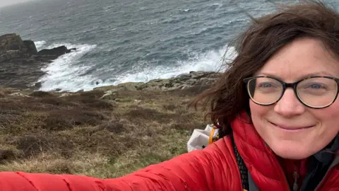 Claire Allen A selfie of Claire Allen wearing a red coat and glasses. Behind her is the sea, bracken and heather. It is a windy day as her hair is blowing and the sea looks rough with lots of sea foam and spray