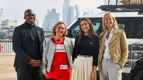 British Red Cross Paul Amadi, chief supporter officer at British Red Cross, Dame Claire Bertschinger, Mehzebin Adam-Suter, curator of Britain's smallest museum and Laura Wood from Jaguar Land Rover are standing next to each other in front of a land rover in London. Ms Bertschinger is wearing a British Red Cross tshirt and is smiling.