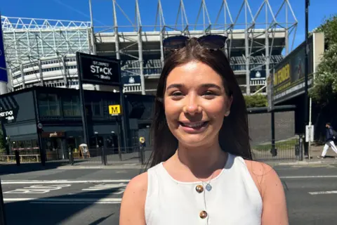 Rachel Riley. She has brown hair and is wearing a white top. She is standing in front of St James' Park.