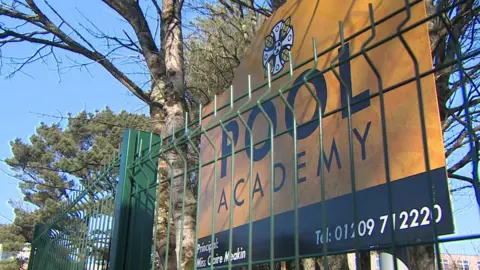 The image shows a sign for Pool Academy behind a green metal fence, with trees and school buildings visible in the background. It appears to be a daytime exterior shot of the school's entrance on a clear, sunny day.