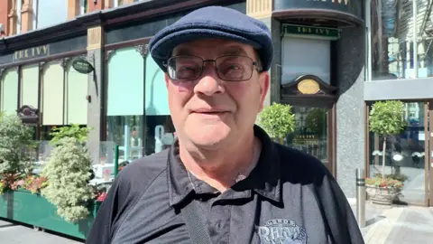 Fiona Callow/BBC A man smiling looking at the camera stood in front of a city centre restaurant. He is wearing a flat cap and a blue T-shirt which has Leeds Rhinos written on. 