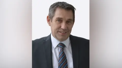 Norfolk and Norwich University Hospital Chief operating officer, Chris Cobb looks at the camer in a black suit and white shirt He is wearing a striped blue and orange tie. His hair is swept to one side and he is smiling at the camera