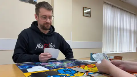 Photograph of Hughes mid-game. He's holding a hand of cards, and is looking down towards the table in front of him, which has brightly coloured playing mats. He's wearing a black hoodie with a white and red stylised design on the front. He's indoors, in a room with cream walls and a window.