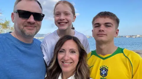 Sharon Paton A family of four, mum, dad and a teenage boy and girl, pose in front of the sea on a sunny day. The man is wearing sun glasses and a blue T-shirt. The woman has long wavy brown hair with blonde highlights. The brown-haired boy has a yellow and green Brazil football shirt. The girl is fair and is wearing a white T-shirt. All are smiling.