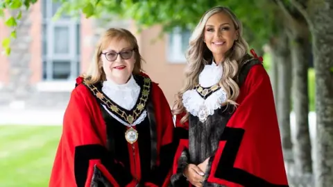 Antrim and Newtownabbey Council Deputy Mayor Julie Gilmour and Mayor Leah Kirkpatrick pose together wearing red ceremonial robes and their gold chains of office. Kirkpatrick has long, wavy blonde hair. Gilmour has shoulder-length blonde hair with a fringe and is wearing dark glasses. Both women are smiling, there are trees and a lawn in the background.
