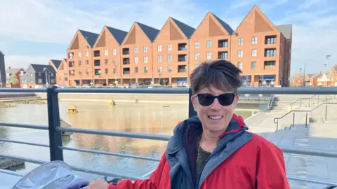 Amy Holmes/BBC A picture of a woman with short black hair who is wearing sunglasses. She is stood in front of a body of water that has six houses with triangle roofs behind it. You can see the top of the pram she is pushing.