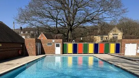 An image showing an outdoor swimming pool on a sunny day. The image is taken from one end looking down to the other. At the far end are changing rooms with green, yellow, dark blue and red doors. Behind them is a large tree with no leaves, with houses in the distance.
