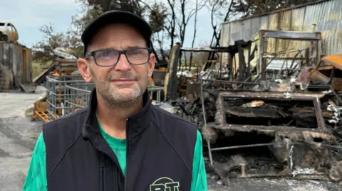 John Harvey stands in the destroyed yard of his MOT Centre. There are charred vehicles around him. He's wearing a dark cap, glasses. and a black gilet. 
