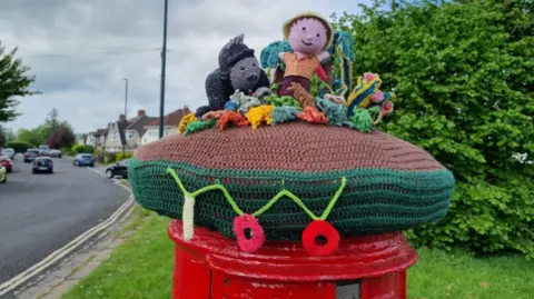 Lynne Harrison A postbox with a knitted topper featuring Sir David and a gorilla sitting among multicoloured woollen plants. There is a residential road stretching off to the left and a hedge to the right.