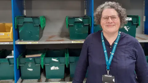 Vicky Fraser Vicky is stood in front of shelves of green stash bins used to help sort books and stock that come in to be distributed to libraries. She has long dark curly hair, is wearing glasses and a dark blue top. She is wearing a blue lanyard and is smiling at the camera.