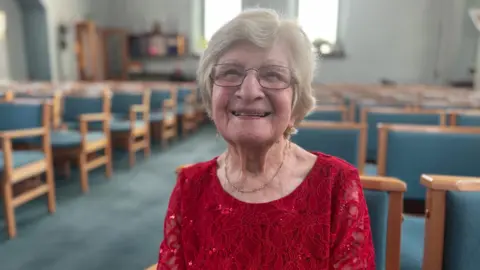 An elderly lady with white hair and glasses, wearing a red dress inside Long Eaton United Reformed Church