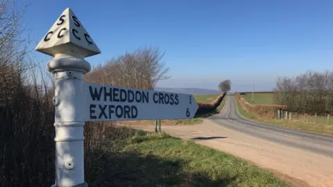 View in the background of a sunny road. There are blue skies in the distance and hills. The signpost in the foreground is a white fingerpost sign which reads "Wheddon Cross 3/4 and Exford 6".