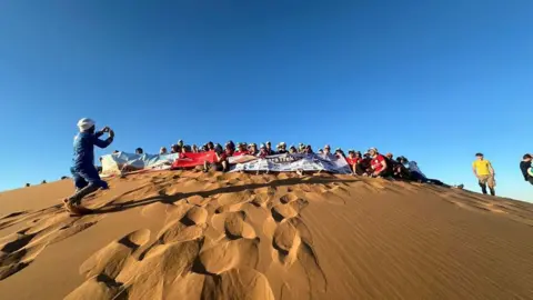 Family A large group of people at the top of a large sand dune with footprints in the sand leading to the top