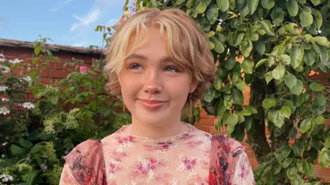 Close up of Niamh Shergold. She is stood outside in front of a red brick wall with green trees and shrubbery. She has short light hair and is wearing a pink floral top.