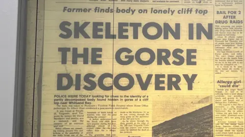 Image shows a section of a newspaper page lit up by a microfilm machine. Part of the machine is visible on the left hand side of the frame. The headline of the story is: Farmer finds body on lonely cliff top: Skeleton in the gorse discovery. There is also a black and white picture of part of the clifftop with the sea and another peninsula in the background