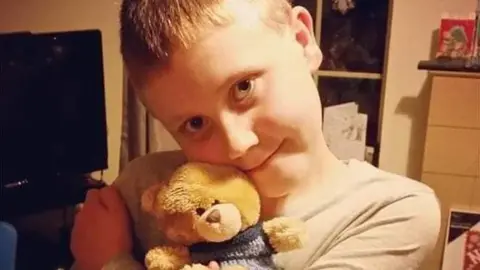 A young boy clutching a teddy is looking at the camera. The boy has short, light coloured hair 