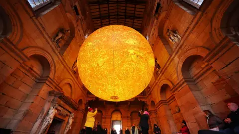 Visitors stand under Helios and take photographs of the sculpture at Seaton Delaval Hall. The Sun artwork is hanging in the old building, surrounded by stone pillars and sculptures.