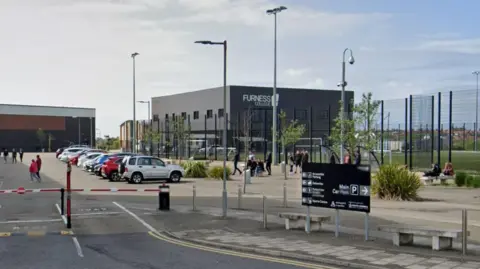 Furness College. A grey cube building stands behind a number of football pitches on the college campus. There are parked cars and people milling about. A dark grey, large rectangular sign in the foreground shows directions to various buildings. There are two thin red and white pole barriers at the entrance to the parking area.