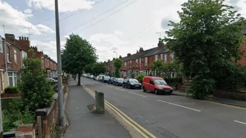Google A residential street lined with rows of red‑brick terraced houses on both sides. Several parked cars sit along the left side of the road, including a prominent red van on the right. A few trees and small front gardens border the pavement. The sky is partly cloudy, and the road has double yellow lines along the edges of both pavements.