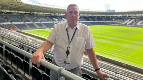 BBC A man with grey hair and wearing a white shirt, grey trousers and black lanyard  stands among black seats in a large sports stadium with his hand on a steel rail. A grass pitch can be seen in the background.