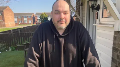 Shot of Delboy Booth leaning on a fence outside his house. He has short hair and a beard and is wearing a black top. 