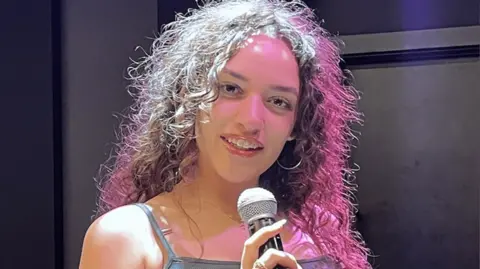 Iquao Aluko in a music studio at the college holding a microphone. She has curly hair and wears a blue dress and hoop earrings.