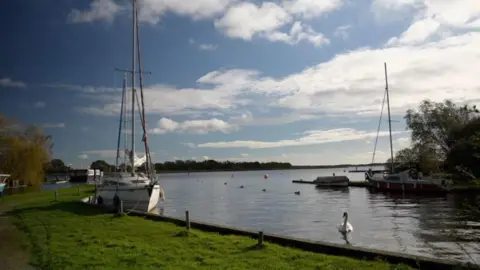 Hickling Broad has white-bodied sailing boats moored on the banks and a swan swimming in the water.