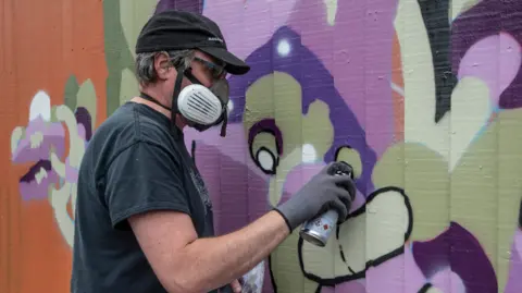 Andy Council Andy Council wearing a grey t-shirt and gloves, spraying a purple and grey/beige mural on a wall with a spray can. He is wearing a white mask aqnd a black cap.