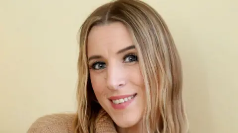 A photo of Faye Louise. She is smiling at the camera, is wearing a brown wooly turtle neck style jumper and has blonde hair. She is smiling.