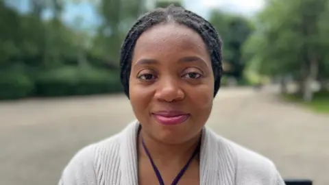 Perpetua has brown skin and braided black hair, tied back. She is smiling at the camera and is wearing a strappy, halter-neck purple top, with a cream cardigan on top. She is sat down and behind her, blurred, is an open concrete pathway and trees.