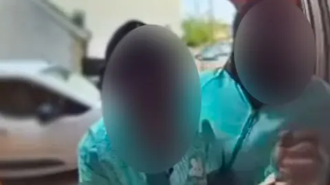 Two men are wearing turquoise Deliveroo branded jackets, and are leaning towards the camera, but their faces are blurred out. Behind them is a white car and a street scene, but the background is out of focus.