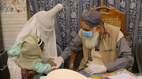 A man sitting in a chair wearing a stethoscope, face mask, and gloves greets a woman holding her child. The woman and child's faces are obscured.